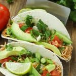 Two taco shells filled with slow cooker taco chicken and topped with avocado, tomato and green onions.
